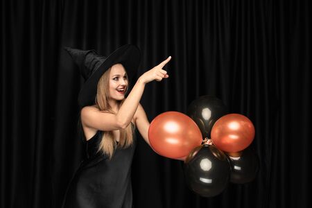 Young blonde woman in black hat and costume on black background. Attractive, sensual female model. Halloween, black friday, cyber monday, sales, autumn. Copyspace. Holding balloons, pointing, smiling.の写真素材