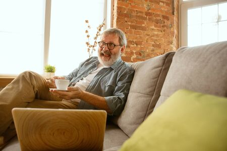 Senior man working with laptop at home - concept of home studying. Caucasian male model sitting on sofa and having coffee break while serfing in internet, watching cinema or webinar, online lessons.の写真素材