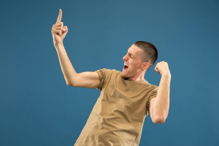 Caucasian young mans half-length portrait on blue studio background. Beautiful male model in shirt. Concept of human emotions, facial expression, sales, ad. Celebrating, crazy happy, screaming.の写真素材