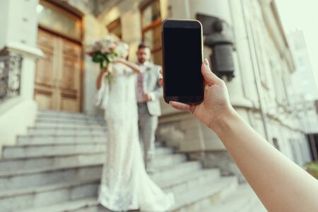Guest making photo of caucasian romantic young couple celebrating marriage in city. Bride and groom. Family, relation, love concept. Contemporary wedding. Moments. Memories of important day.