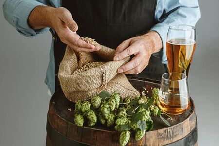 Close up of confident senior man brewer with self crafted beer in glass on wooden barrel on grey...