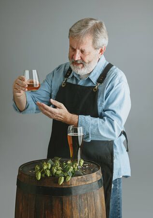 Confident senior man brewer with self crafted beer in glass on wooden barrel on grey background....