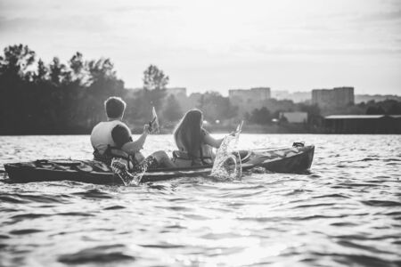 Happy young caucasian couple kayaking on river with sunset in the backgrounds. Having fun in leisure activity. Happy male and female model laughting on the kayak. Sport, relations concept. Colorless.の写真素材