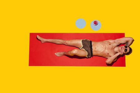 Top view of young caucasian male models resting on beach resort on red mat and yellow background. Mans lying under sunlight and talking on phone. Concept of summertime, party, chill, vacation.の写真素材