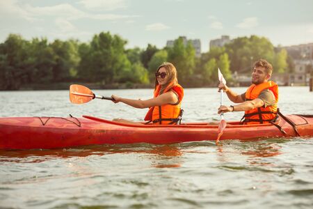 Happy young caucasian couple kayaking on river with sunset in the backgrounds. Having fun in leisure activity. Happy male and female model laughting on the kayak. Sport, relations concept. Colorful.の写真素材