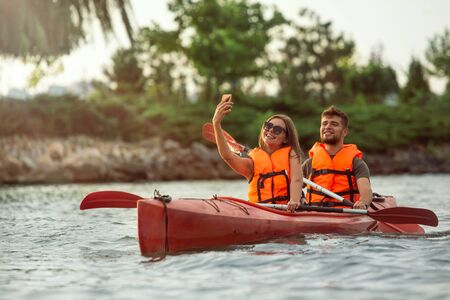 Happy young caucasian couple kayaking on river with sunset in the backgrounds. Having fun in leisure activity. Happy male and female model laughting on the kayak. Sport, relations concept. Colorful.の写真素材