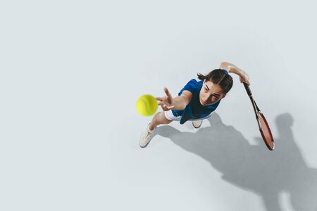 Young woman in blue shirt playing tennis. She hits the ball with a racket. Indoor studio shot isolated on white. Youth, flexibility, power and energy. Negative space. Top view.の写真素材