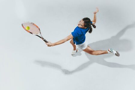 Young woman in blue shirt playing tennis. She hits the ball with a racket. Indoor studio shot isolated on white. Youth, flexibility, power and energy. Negative space. Top view.の写真素材