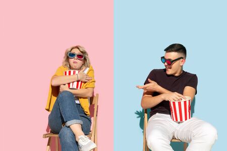 Young emotional man and woman in bright casual clothes posing on pink and blue background. Concept of human emotions, facial expession, relations, ad. Beautiful couple watching cinema with popcorn.の写真素材