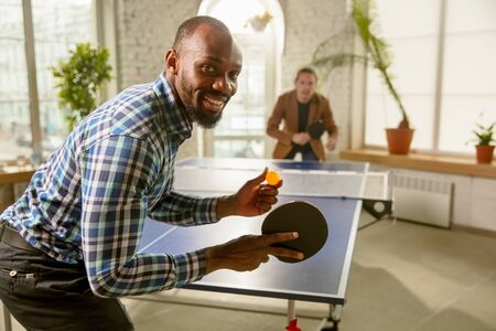 Young men playing table tennis in workplace, having fun. Friends in casual clothes play table tennis together at sunny day. Concept of leisure activity, sport, friendship, team-building, teamwork.の写真素材