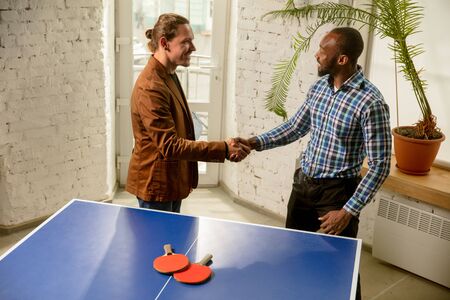 Young men playing table tennis in workplace, having fun. Friends in casual clothes play table tennis together at sunny day. Concept of leisure activity, sport, friendship, team-building, teamwork.の写真素材