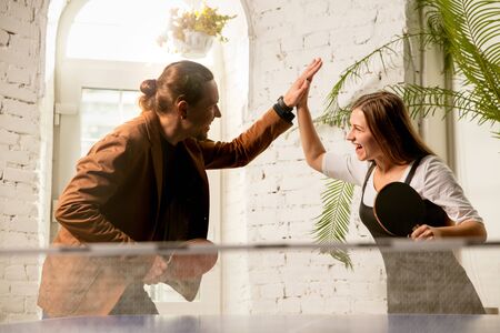 Young people playing table tennis in workplace, having fun. Friends in casual clothes play table tennis together at sunny day. Concept of leisure activity, sport, friendship, team-building, teamwork.の写真素材