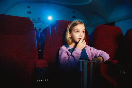 Little caucasian girl watching a film at a movie theater, house or cinema. Looks expressive and emotional. Sitting alone and having fun. Holidays, leisure activity, childhood, weekend.の写真素材