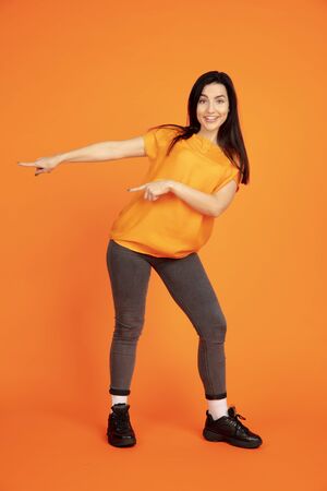 Caucasian young womans portrait on orange studio background. Beautiful female brunette model in shirt. Concept of human emotions, facial expression, sales, ad. Copyspace. Pointing, showing.の写真素材