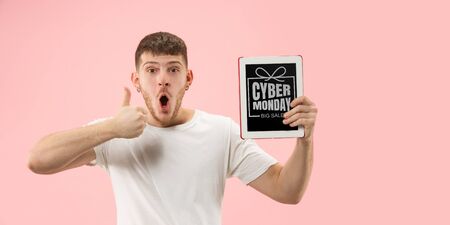 Portrait of young man showing tablet screen on coral background. Looks astonished, shocked. Concept of sales, black friday, cyber monday, finance, business. Online shops and payments bill.の写真素材