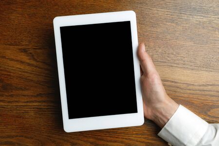 Male hand holding tablet with empty screen on wooden background for text or design. Blank gadget...