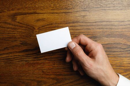 Male hand holding a blank business card on wooden background for text or design. Blank credit card templates for contact or use in business. Finance, office. Mock up. Copyspace.の写真素材