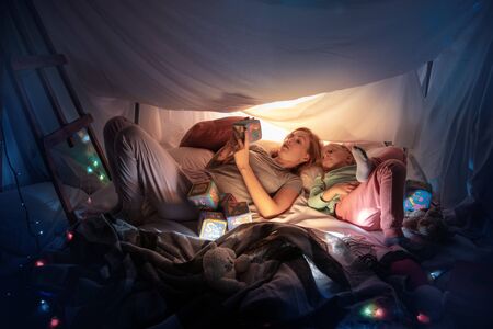 Mother and daughter sitting in a teepee, having fun, playing with the flashlight in dark room with toys and pillows. Look happy. Home comfort, family, love, Christmas holidays, storytelling time.の写真素材