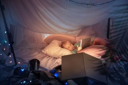 Little girl lying in a teepee, having fun, playing with the flashlight in dark room with toys and...