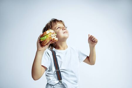Pretty young curly boy in casual clothes on white studio background. Eating burger. Caucasian male preschooler with bright facial emotions. Childhood, expression, fun, fast food. Dreamful looks up.の写真素材