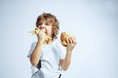 Pretty young curly boy in casual clothes on white studio background. Eating burger with fried potato. Caucasian male preschooler with bright facial emotions. Childhood, expression, fun, fast food.の写真素材