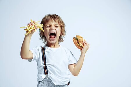 Pretty young curly boy in casual clothes on white studio background. Eating burger with fried potato. Caucasian male preschooler with bright facial emotions. Childhood, expression, fun, fast food.の写真素材