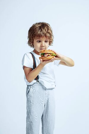 Pretty young curly boy in casual clothes on white studio background. Eating burger. Caucasian male preschooler with bright facial emotions. Childhood, expression, having fun, fast food. Astonished.の写真素材