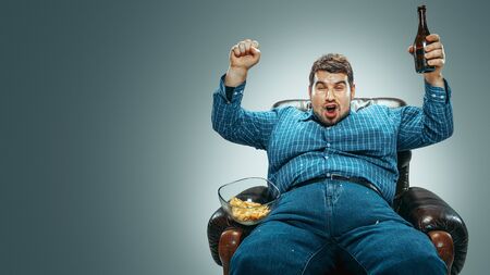 Portrait of fat caucasian man wearing jeanse and whirt sitting in a brown armchair isolated on gradient grey background. Watching TV drinks beer, eats chips and changing channels. Overweight, carefree.の写真素材
