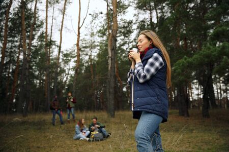 Close up of woman drinking coffee or tea with group of friends on a hiking trip in autumn day....