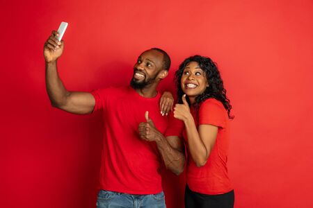 Young emotional african-american man and woman in bright casual clothes posing on red background. Concept of human emotions, facial expession, relations, ad. Couple making selfie, pointing, smiling.の写真素材