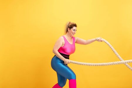 Young caucasian plus size female models training on yellow background. Copyspace. Concept of sport, healthy lifestyle, body positive, fashion, style. Stylish woman practicing with ropes and smiling.の写真素材
