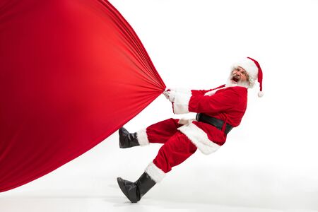 Santa Claus pulling huge bag full of christmas presents isolated on white background. Caucasian male model in traditional costume. New Year 2020, gifts, holidays, winter mood. Copyspace for your ad.の写真素材