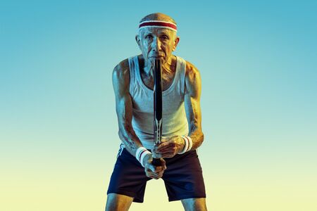 Senior man wearing sportwear playing tennis on gradient background, neon light. Caucasian male model in great shape stays active, sportive. Concept of sport, activity, movement, wellbeing, confidence.の写真素材