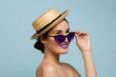 Portrait of beautiful woman with bright make-up, hat and sunglasses on blue studio background. Stylish and fashionable make and hairstyle. Colors of summer. Beauty, fashion and ad concept. Retro.の写真素材