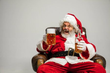 Santa Claus drinking beer sitting on armchair, congratulating, looks drunk and happy. Caucasian male model in traditional costume. New Year 2020, gifts, holidays, winter mood. Copyspace for your ad.の写真素材