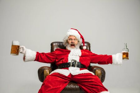 Santa Claus drinking beer sitting on armchair, congratulating, looks drunk and happy. Caucasian male model in traditional costume. New Year 2020, gifts, holidays, winter mood. Copyspace for your ad.の写真素材