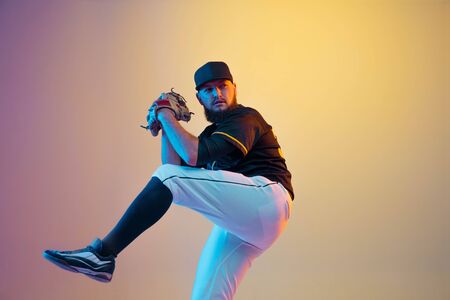 Baseball player, pitcher in a black uniform practicing and training on gradient background in neon light. Young professional sportsman in action and motion. Healthy lifestyle, sport, movement concept.の写真素材