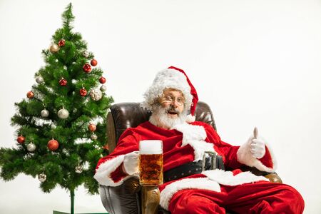 Santa Claus drinking beer near the Christmas tree, congratulating, looks drunk and happy. Caucasian male model in traditional costume. New Year 2020, gifts, holidays, winter mood. Copyspace for your ad.の写真素材