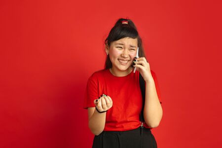 Asian teenagers portrait isolated on red studio background. Beautiful female brunette model with long hair in casual style. Concept of human emotions, facial expression, sales, ad. Talking on phone.の写真素材