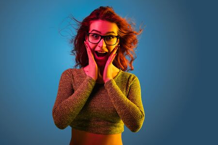 Caucasian womans portrait isolated on blue studio background in neon light. Beautiful female model with red hair in casual style. Concept of human emotions, facial expression, sales, ad. Astonished.の写真素材