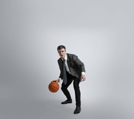 Back in college - its never too late for being sport star. Man in office clothes traines in basketball on grey background. Unusual look for businessman in motion, action. Sport, healthy lifestyle.の写真素材