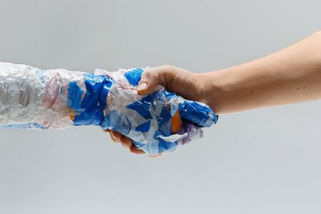 Big plastic hand made of garbage shaking another hand on white studio background. Polymers overusing...