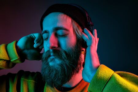 Calm, happy. Caucasian mans portrait on gradient studio background in neon light. Beautiful male model with hipster style in earphones. Concept of human emotions, facial expression, sales, ad.の写真素材