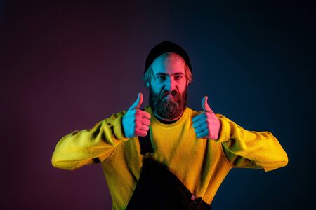 Confident showing thumbs up. Caucasian man portrait on gradient studio background in neon light. male model with hipster style.の写真素材