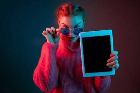 Showing blank tablet screen. Caucasian woman's portrait isolated on gradient studio background in neon light. Beautiful female model with red hair. Concept of human emotions, facial expression, ad.の写真素材