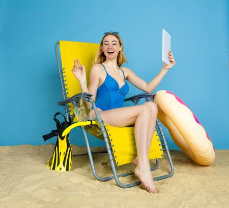 Happy young woman with tablet takes selfie or vlog about traveling on blue studio background. Concept of human emotions, facial expression, summer holidays, weekend. Summertime, sea, ocean, alcohol.の写真素材