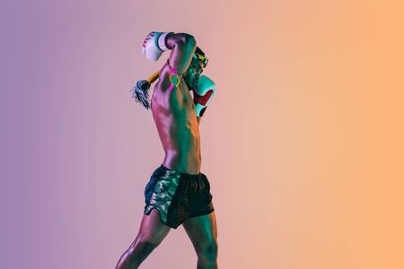 Muay thai. Young man exercising thai boxing on gradient background in neon light. Fighter practicing, training in martial arts in action, motion. Healthy lifestyle, sport, asian culture concept.の写真素材