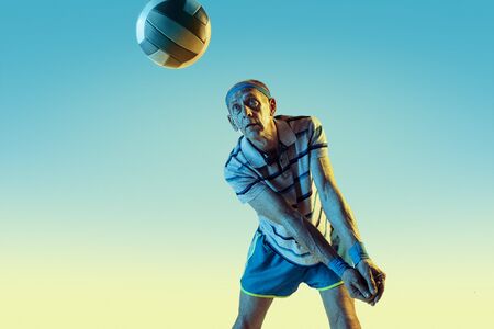 Senior man wearing sportwear playing volleyball on gradient background, neon light. Caucasian male model in great shape stays active. Concept of sport, activity, movement, wellbeing, confidence.の写真素材