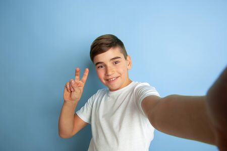 Taking selfie with sign of peace. Caucasian boy portrait isolated on blue studio background....