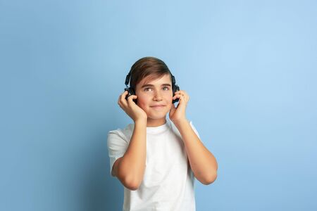 Listen to music, looks joyful. Caucasian boy portrait isolated on blue studio background. Beautiful teen male model in white shirt posing. Concept of human emotions, facial expression, sales, ad.の写真素材